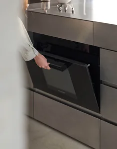 A minimalist kitchen with a metallic oven door being opened by a person with light-colored hair.