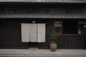 A traditional Japanese storefront with wooden shutters, concrete steps, and a large fabric curtain. A small plant in a pot ad