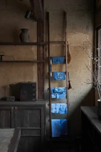 A wooden ladder with tied-dye towels hanging on it in a rustic interior space with natural lighting and a vase on a shelf.