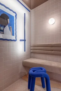 A minimalist restaurant interior featuring ceramic tiles, a bench, and a blue stool under a wall-mounted light.