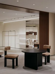 A modern retail interior showcases a round, black table and beige chairs, surrounded by white shelves and concrete walls.