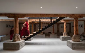 Monks ascending a black staircase in a room with wooden beams and stone sculptures, featuring a modern architectural design.