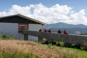 Four monks in maroon robes standing on a concrete bridge over wild grasses toward a modern, white-walled monastery building i