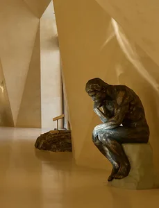 A bronze sculpture of a seated figure with crossed legs, sitting in a thought-provoking pose, placed on a marble pedestal in