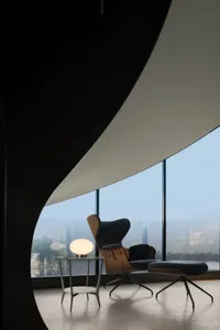 Image of a modern living room with a wooden chair and ottoman, large windows, and a table lamp