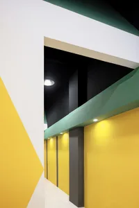A modern interior corridor with geometric lighting featuring a mix of yellow, white, and green walls and concrete pillars.