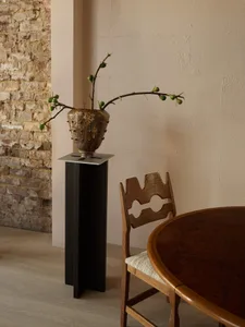 A modern dining room setup featuring a wooden and metal plant stand with a stone vase and greenery, against a textured brick 