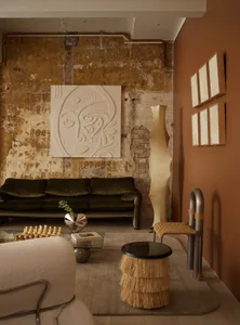 A modern living room with a green velvet sofa, beige textured armchair, and a beige fringed ottoman, featuring a rustic coppe