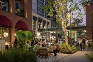 A bustling urban commercial space featuring brick and concrete buildings, lush greenery, and illuminated trees with lights.