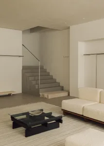 Modern minimalist living room with concrete staircase, wooden furniture, and glass table.