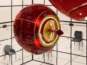 A red metallic sphere with a glowing gold interior, suspended in a modern installation setting.