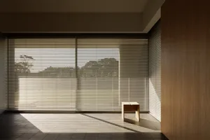 Minimalist interior room with wooden floors and a single wooden bench in front of a large window with blinds.