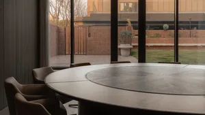 A modern office space featuring a round wooden table and chairs surrounded by large windows overlooking a green courtyard.