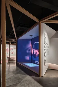 A minimalist exhibition space with a dark interior featuring a large screen displaying a person's face and a wall with the wo