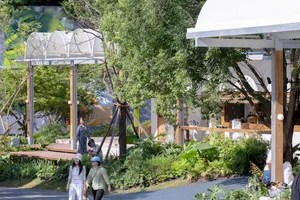 People walking in a modern outdoor area with wooden structures, glass walls, and green plants.