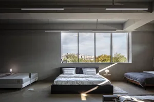 Modern minimalist bedroom with concrete walls, large windows overlooking greenery, and a king-sized bed with white linens.