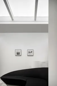 A minimalist living room featuring a sleek black couch on a white floor, with a skylight and framed pictures on the wall.