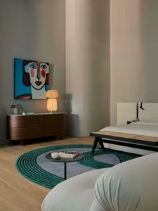 Modern minimalist bedroom featuring a round rug, bed, and side table with lamp and plant.