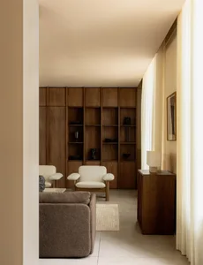 A minimalist living room with neutral tones, featuring a brown sofa, white chairs, and a wooden bookshelf.