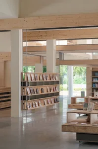 A minimalist modern library with wooden bookshelves, concrete flooring, and large windows allowing natural light.