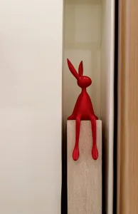A minimalist red plastic sculpture with long legs sitting on a white pedestal in an interior space.