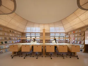 Modern library with wooden furniture and glass walls filled with books, designed for reading and study.