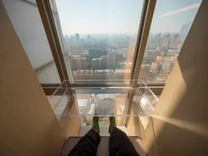 Person's legs seated on a rooftop with a panoramic city view through large glass windows.