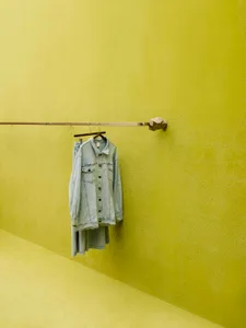 A denim jacket hanging on a metal hanger against a bright yellow wall.