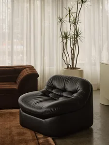 A modern interior featuring a black leather lounge chair, a brown velvet sofa, a tall potted plant, and sheer curtains.