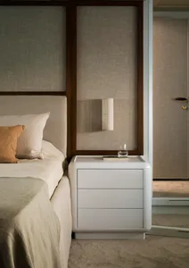 A minimalist bedroom with a white bed and nightstand, featuring a glass and metal lamp and a book.