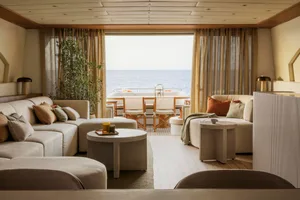 A stylish, modern living room with neutral-toned sofas, ottomans, and tables, featuring large windows with curtains overlooki