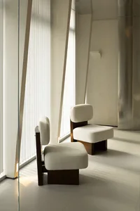 A minimalist living room with white cushions on wooden chairs and stools, featuring vertical blinds and natural light.