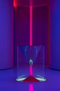 A minimalist installation featuring a large glass cylinder with a conical base, illuminated by a vertical pink light in a cir