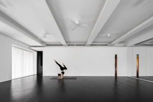 A woman performing a yoga pose in a minimalist studio with white walls and black floor.