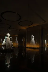 Glass cases displaying elegant dresses in a dimly lit room with circular lights on the ceiling.