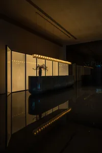 A modern lobby featuring a sleek, black marble reception desk with a vase of dried flowers, illuminated by a linear chandelie
