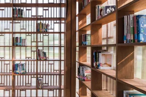 A modern wooden bookshelf filled with books, located in a room with a glass wall providing natural light.
