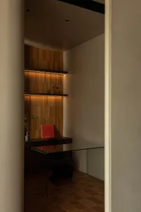 A modern minimalist office with glass desk, red book, black chair, plant, and shelving with books and decorative items.