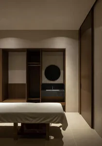A minimalist bedroom featuring a bed with a white sheet, a wooden cabinet, and a sink with a round mirror and black counterto