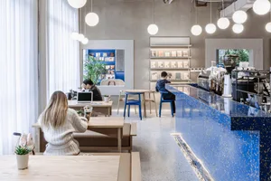 A modern coffee shop with a blue granite counter, white curtains, and potted plants.