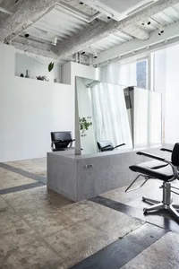 A modern hair salon interior featuring concrete and glass materials, with a black leather salon chair and a large floor-to-ce