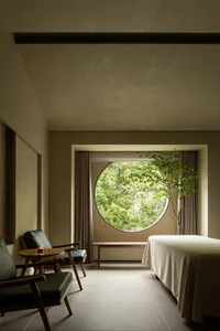 A minimalist bedroom with a round window, textured cloth bedding, and a small tree.