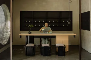 A woman sitting at a wooden table pouring tea, with a shelf of teacups and a bonsai plant.
