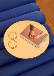 A round, metallic table reflecting jewelry and a book on its surface, placed on a blue, cushioned bench.
