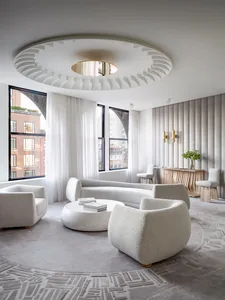 A modern living room with neutral-toned furniture and decor, large windows, and an intricate ceiling design.