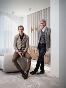 Two men sitting in a modern living room with neutral tones and textured materials.