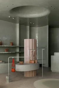 A minimalist retail space features concrete walls and flooring, with bags displayed on a cylindrical pedestal.