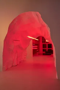 A minimalist interior designed with white plaster walls and a textured archway leading to a modern space with red lighting.
