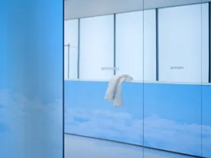 A minimalist office space featuring transparent glass walls and a blue sky and clouds background.