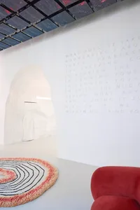 A minimalist white room with a textured white structure, a circular rug with black and pink lines, and a red upholstered chai